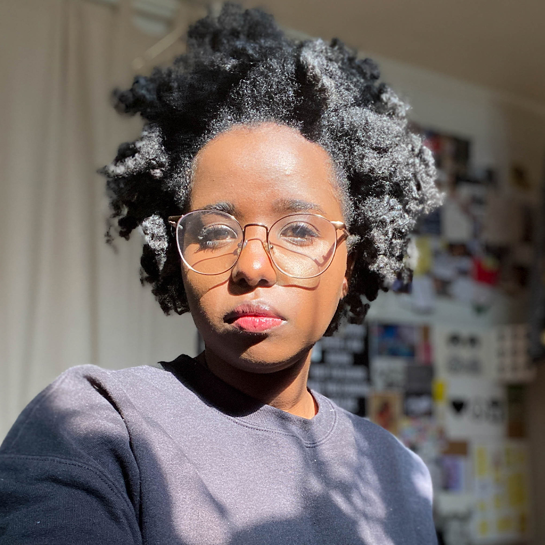 Photo of the artist Enas Satir. She wears glasses and a natural hairstyle while her face glows in the sun. She has a stern expression.