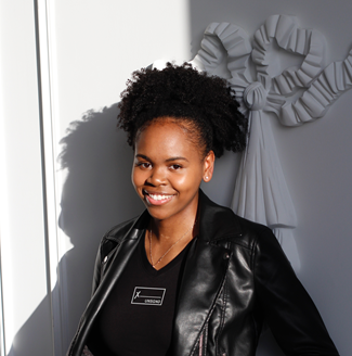 Dymika, a Black woman with her natural hair worn in a high ponytail and wears a cool leather jacket, smiles at the camera in front of a white wall.