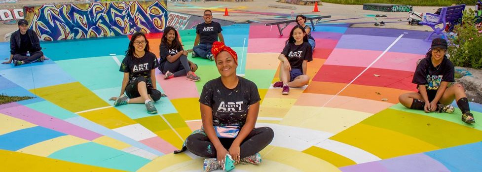 ArtStarts staff sitting on a colourful mural in a semi circle formation. 